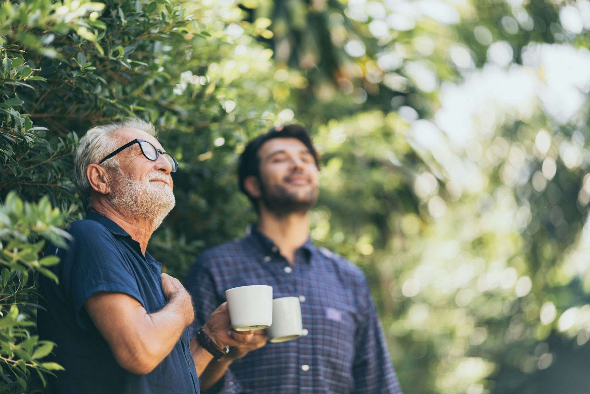 Two men enjoying nature with coffee mugs, surrounded by green foliage, looking content and relaxed.