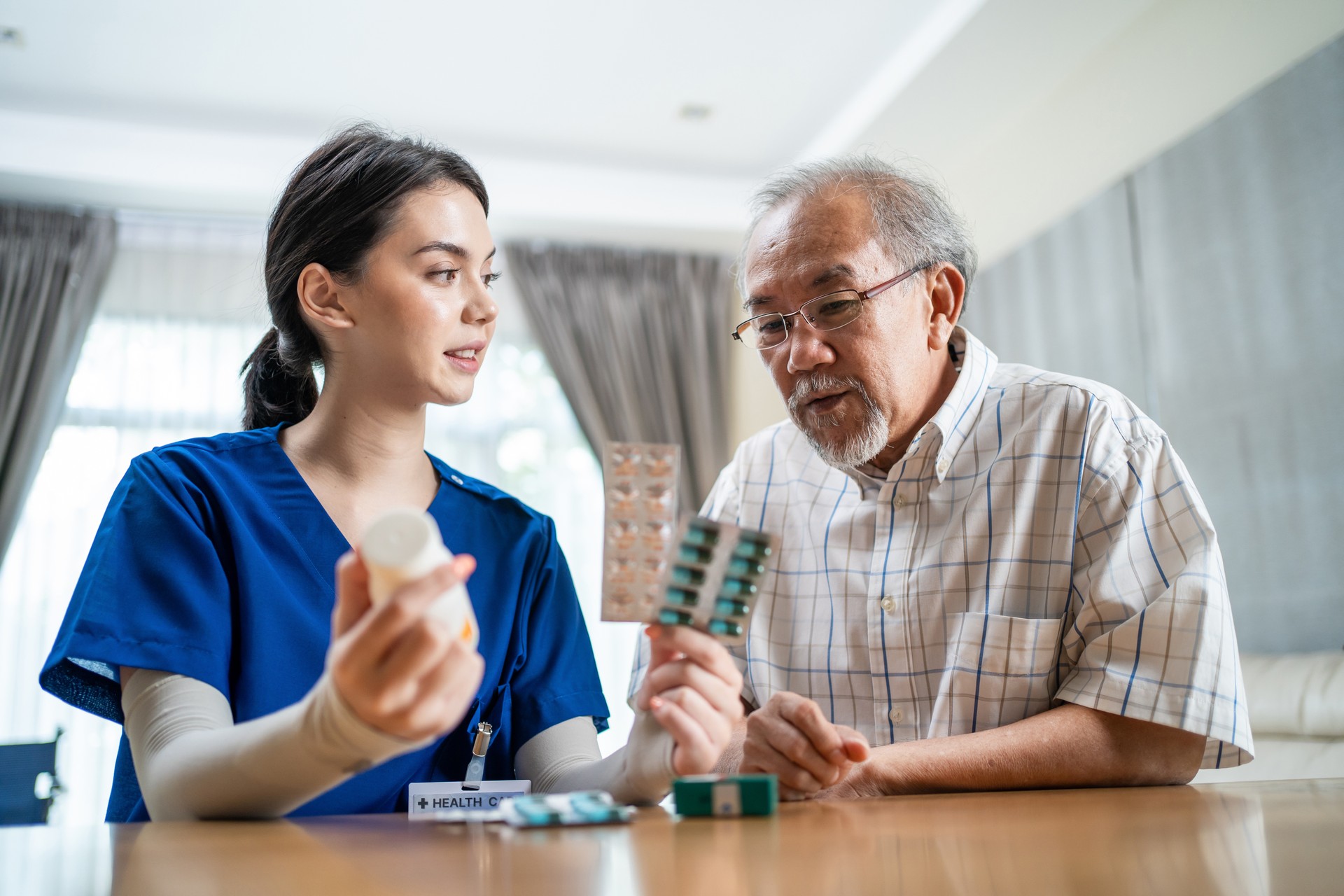 Asian Senior elderly male patient consult with physician nurse at nursing home care. Caregiver therapist pharmacist girl hold medicine pills bottle, explain prescription to older disabled guy in house